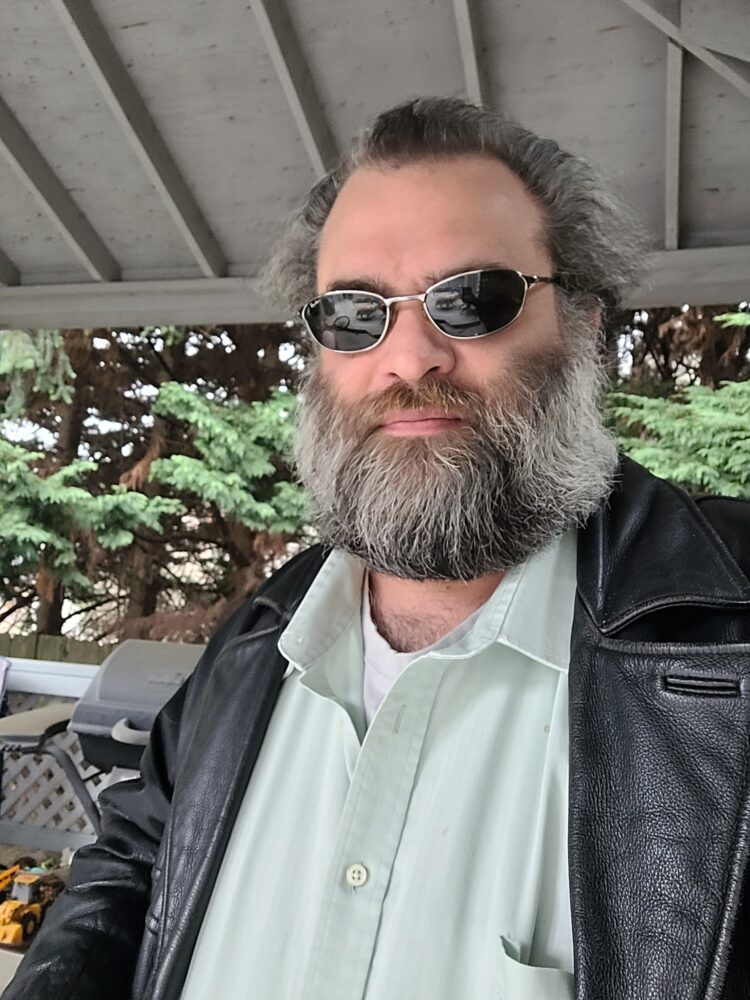 Eli, a middle-aged white man with salt & pepper hair along with full beard, wearing sunglasses, a light green button-up shirt w/collar, and a black leather jacket.